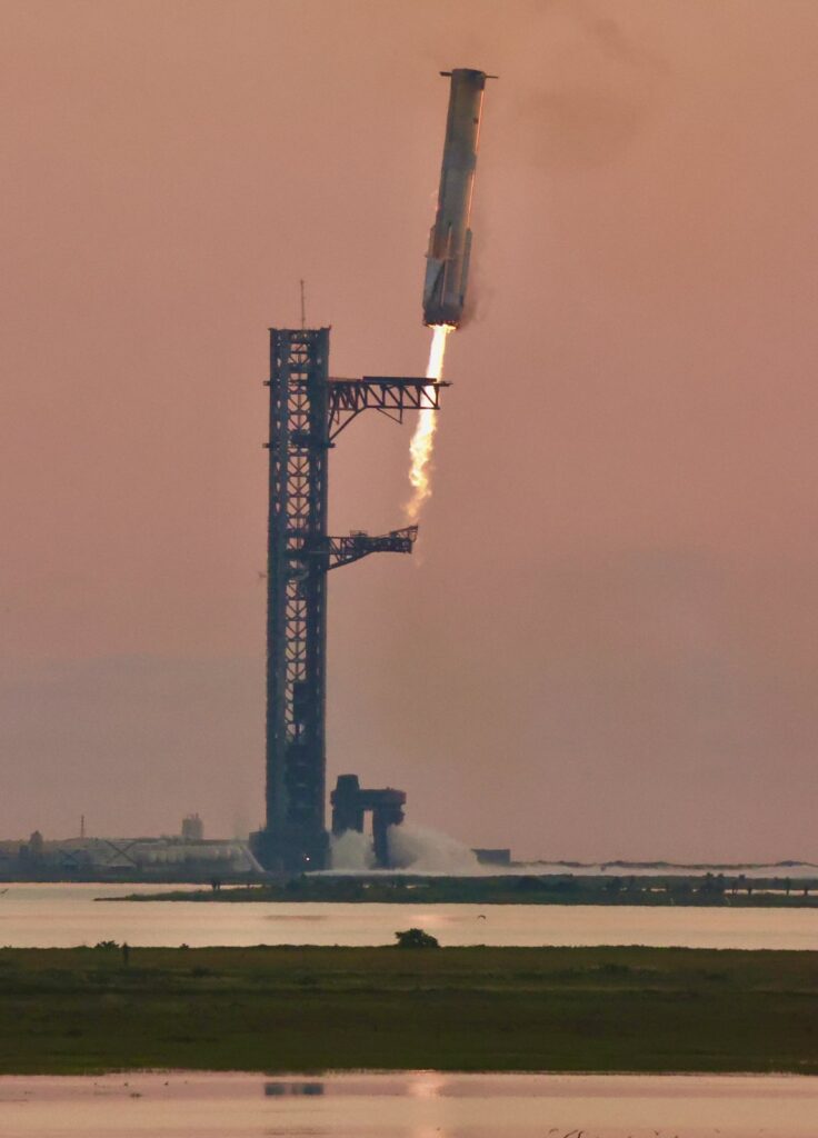 Booster Super Heavy da Starship descendo com os motores acesos ao lado da torre de lançamento, com os braços abertos para a captura, sob céu alaranjado