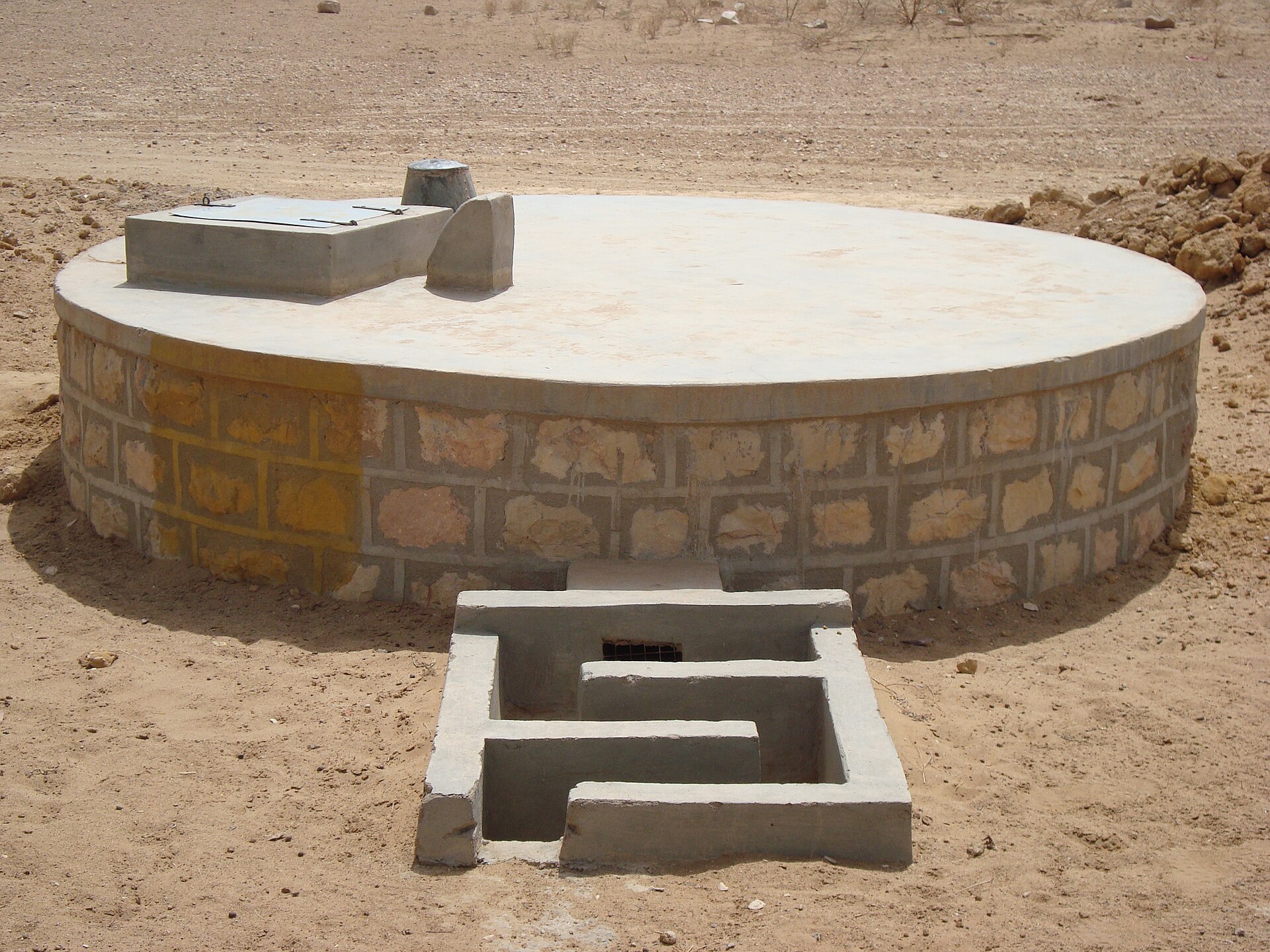 Cisterna redonda de alvenaria rebocada, com tampa de inspeção e canaleta de entrada de água, em terreno de areia