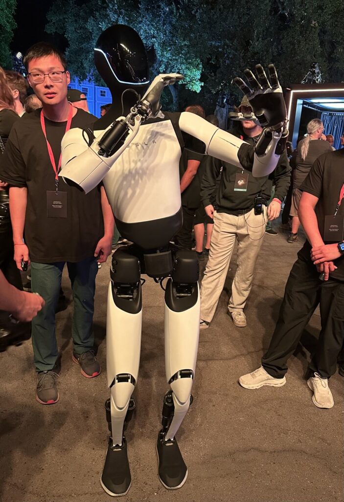 Robô humanoide Optimus, da Tesla, branco e preto, de pé ao ar livre à noite com as mãos levantadas, cercado por pessoas