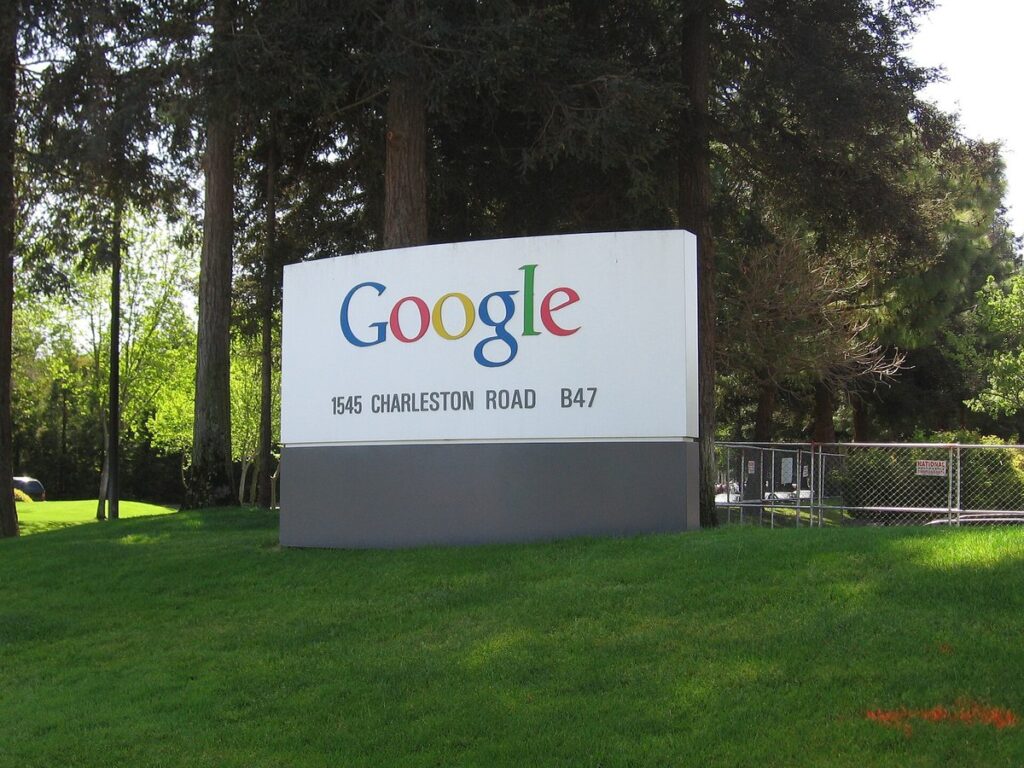 Placa com o logotipo do Google na entrada do Googleplex, em Mountain View, cercada de árvores