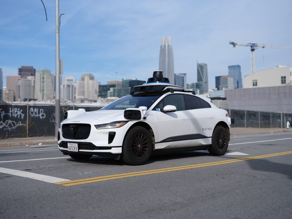 Carro autônomo branco da Waymo, um Jaguar I-Pace com sensores no teto, parado numa rua de São Francisco com prédios ao fundo