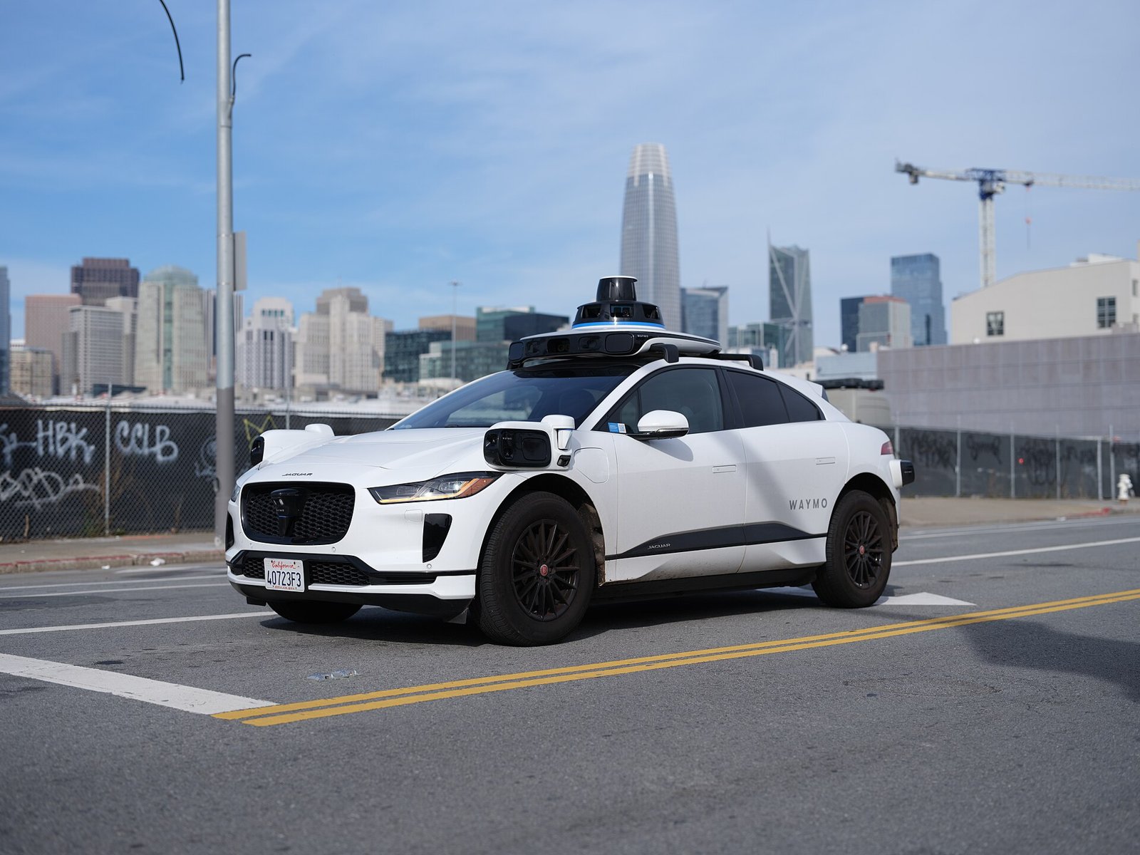 Carro autônomo branco da Waymo, um Jaguar I-Pace com sensores no teto, parado numa rua de São Francisco com prédios ao fundo