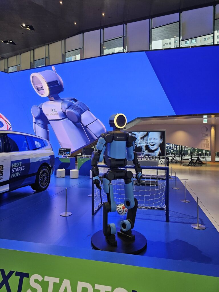 Robô humanoide Atlas, da Boston Dynamics, em pé num palco azul diante de um pequeno gol de futebol, com um telão ao fundo