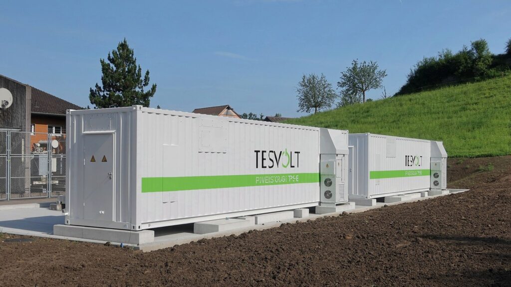 Dois contêineres brancos de baterias da Tesvolt, com faixa verde, instalados ao ar livre sobre uma base de concreto ao lado de um gramado